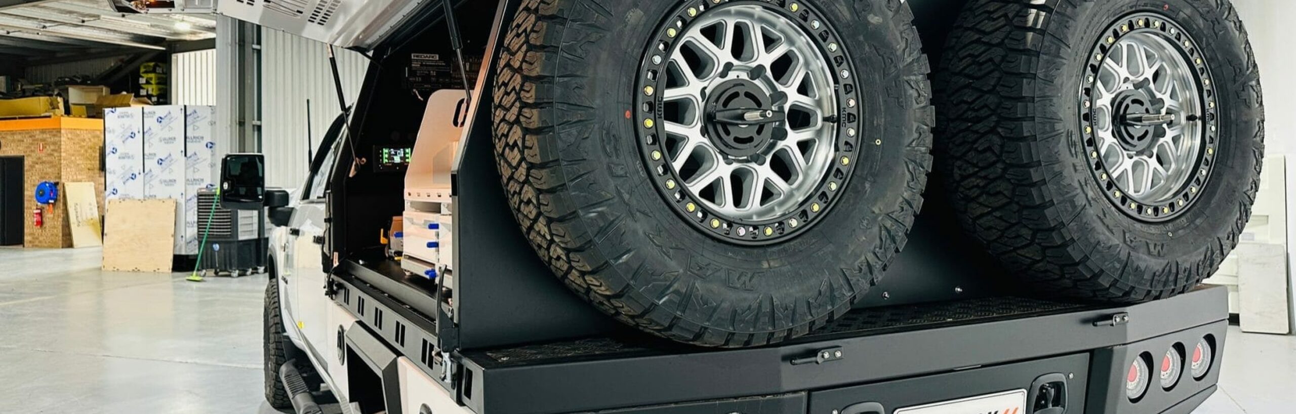 A white dual-cab ute in the OzTrack workshop, fitted with a custom black tray, an open canopy and twin spare wheels mounted at the rear